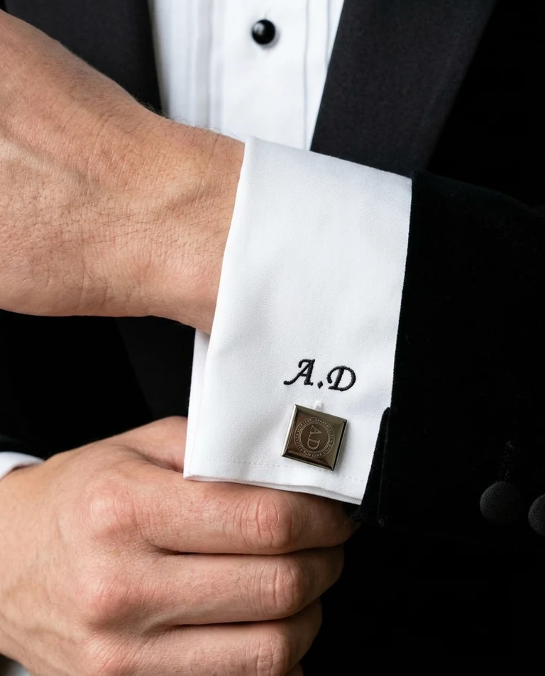 Initials embroidered on a white shirt cuff beside an engraved cufflink, worn with a bespoke black tie dinner suit